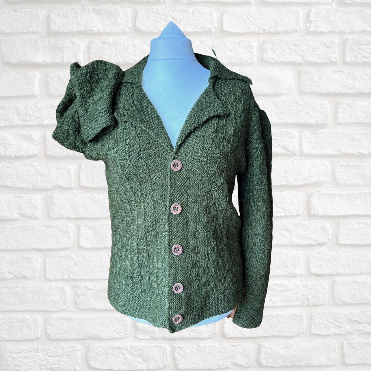 A green textured knit cardigan sweater with ruffled sleeves and pink buttons, displayed on a light blue mannequin torso against a white brick wall background.