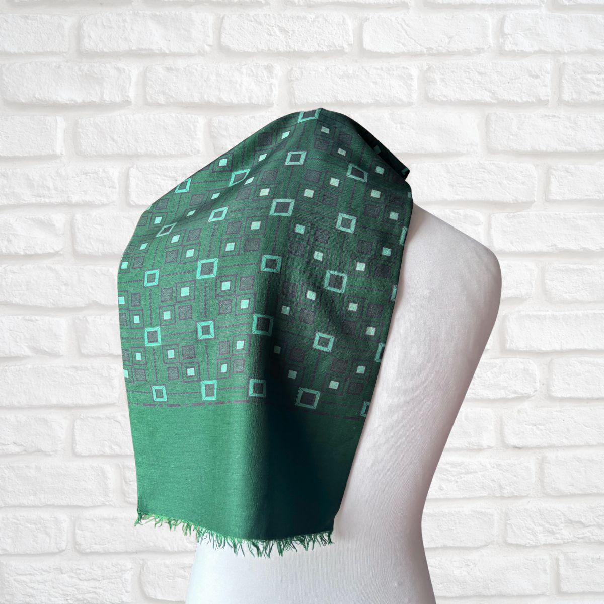 A green patterned scarf draped on a white mannequin torso, set against a white brick wall background.