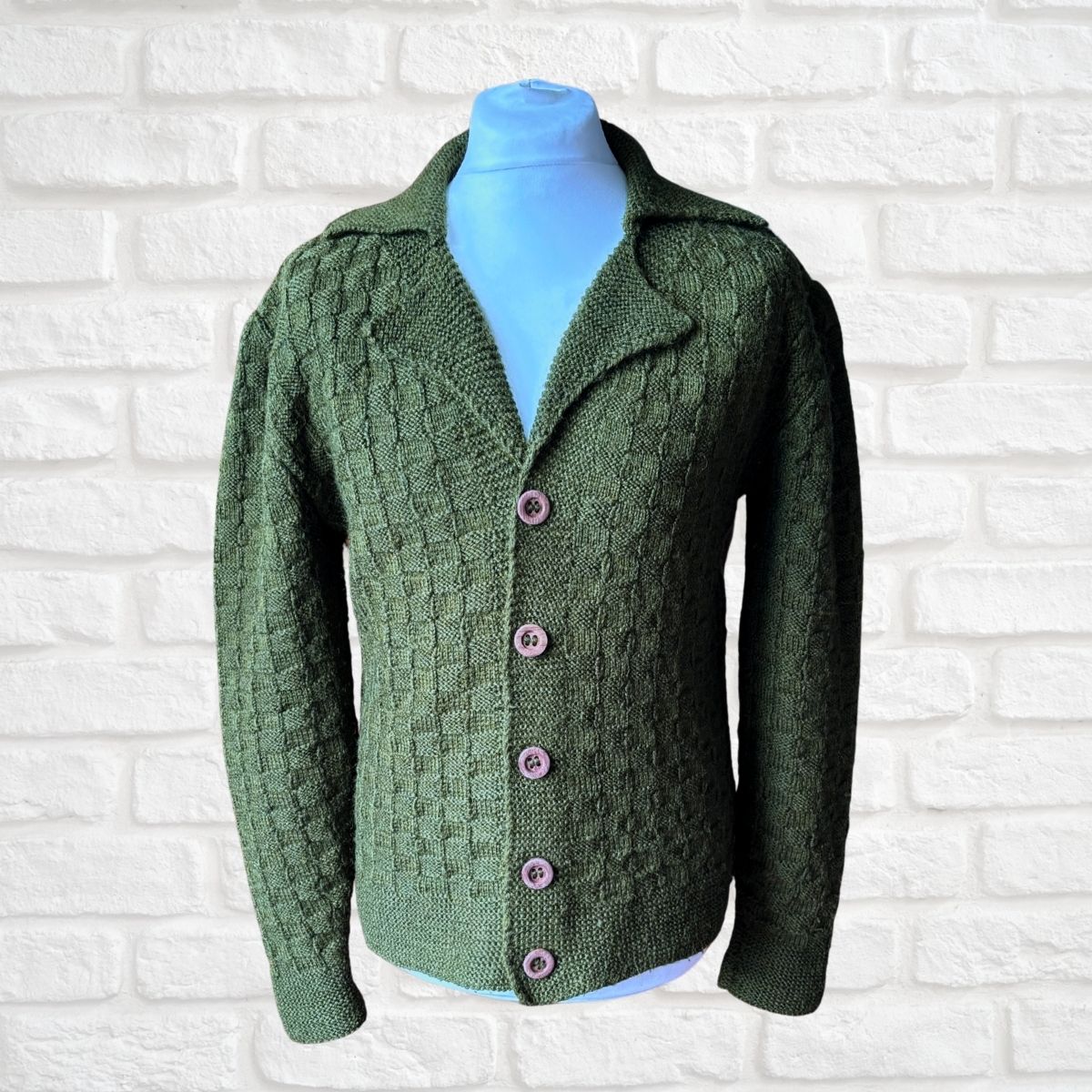 A green cable-knit cardigan sweater displayed on a light blue mannequin torso against a white painted brick wall background.