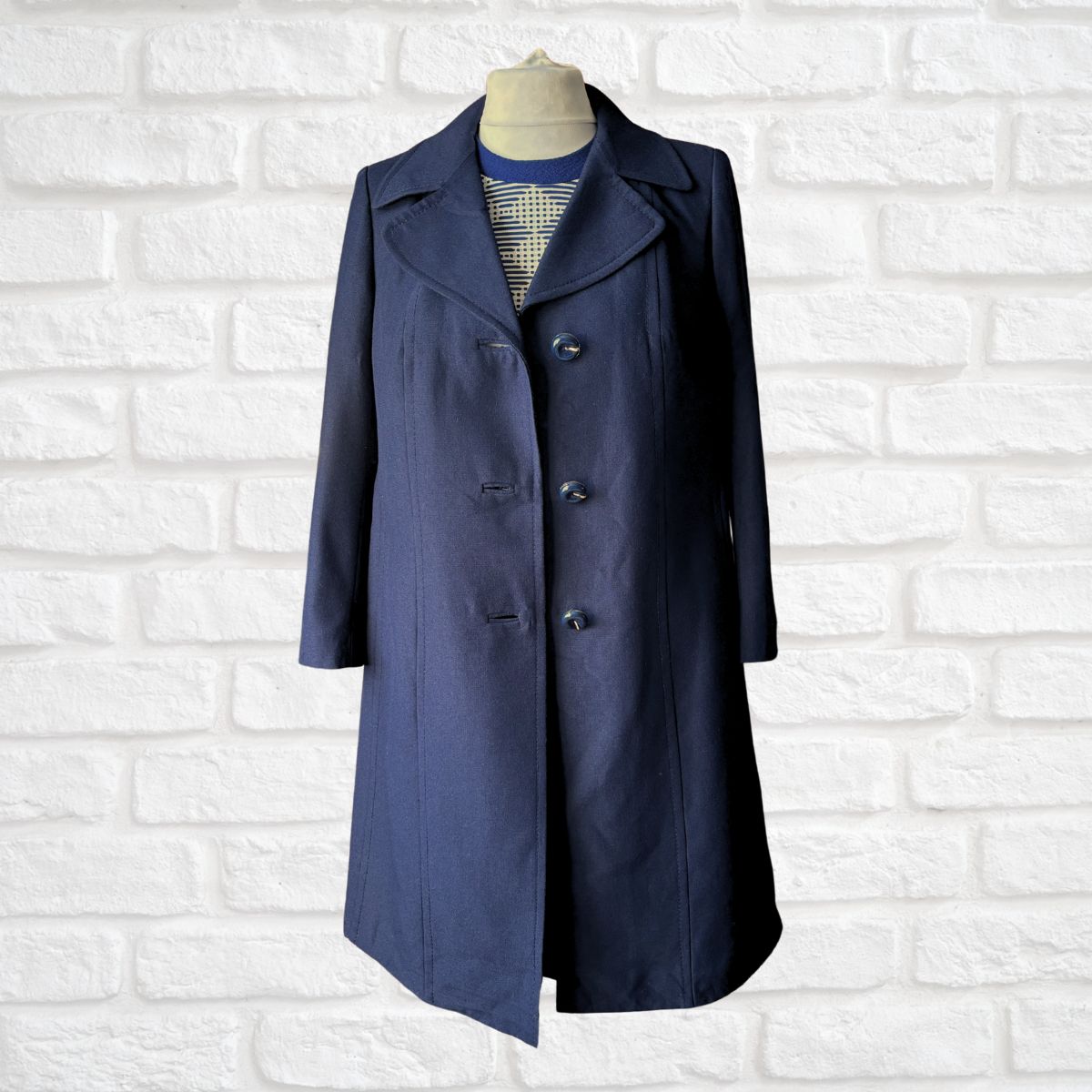 A navy blue mid-length coat displayed on a dress form mannequin against a white painted brick wall background.
