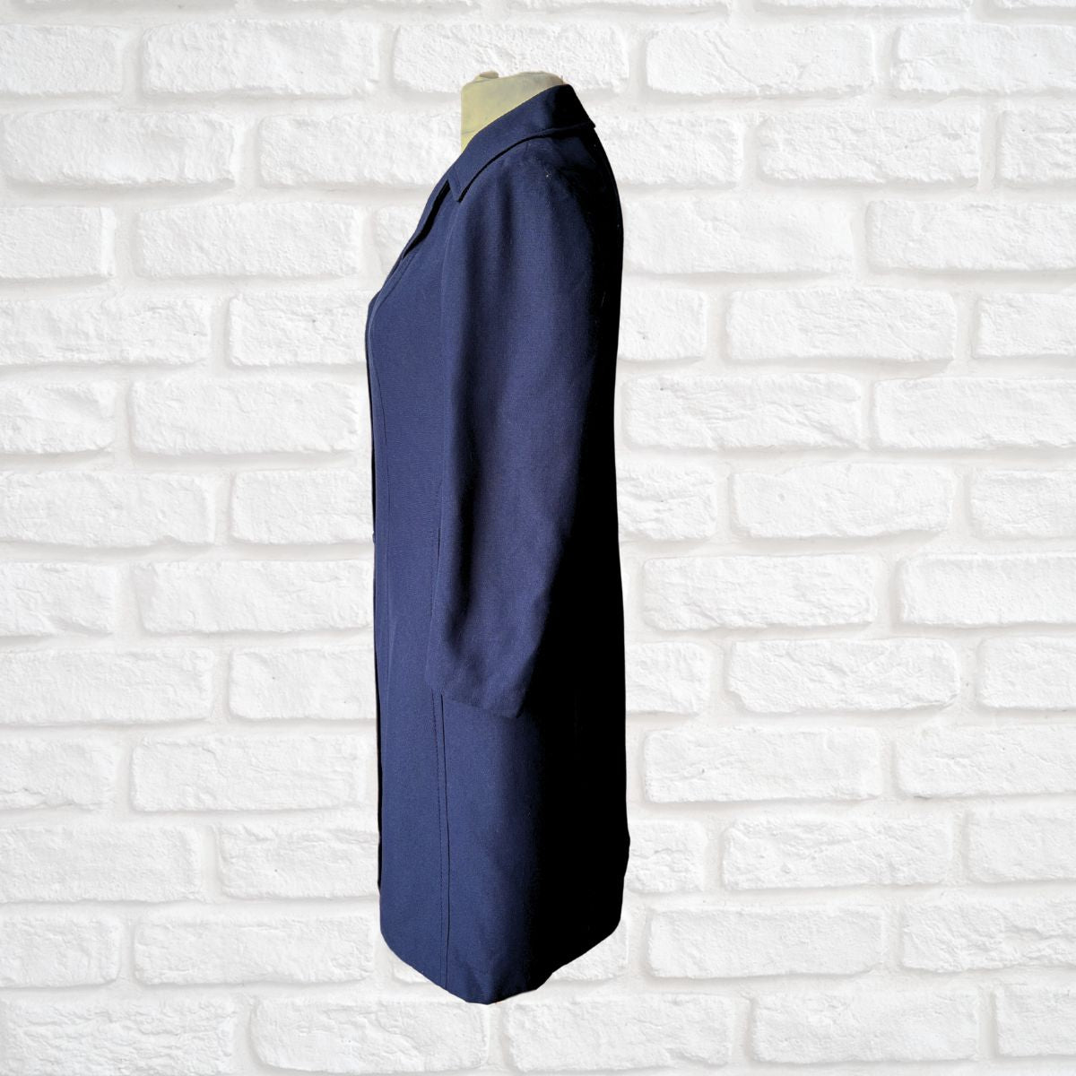 A navy blue long-sleeve coat or jacket displayed on a mannequin torso against a white painted brick wall background.