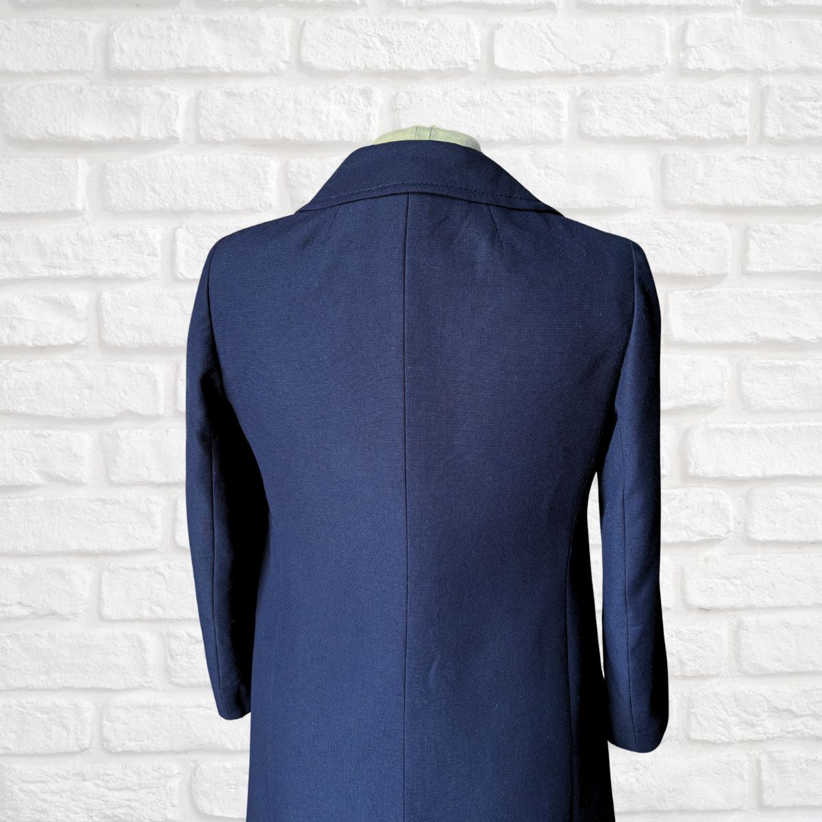 A navy blue women’s coat shown from the back on a mannequin torso, set against a white painted brick wall background.