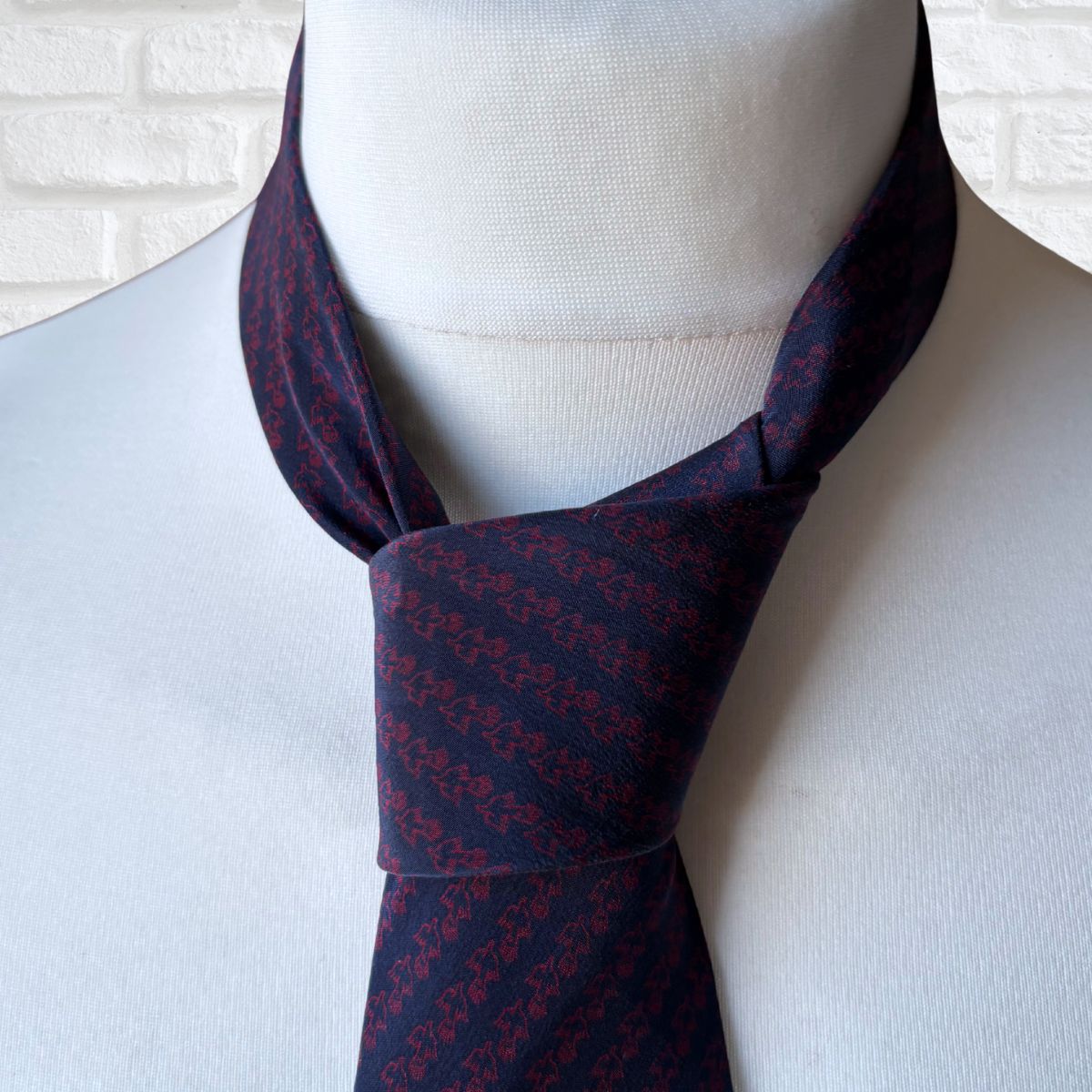 Navy blue necktie with red pattern on a mannequin against a white brick wall.