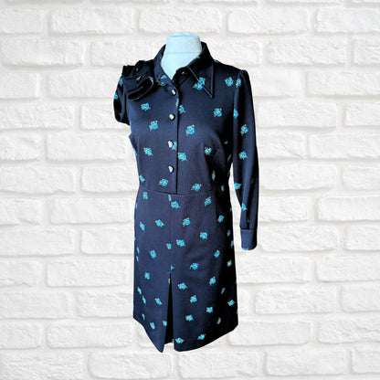 Black dress with blue floral patterns on a mannequin against a white brick wall.