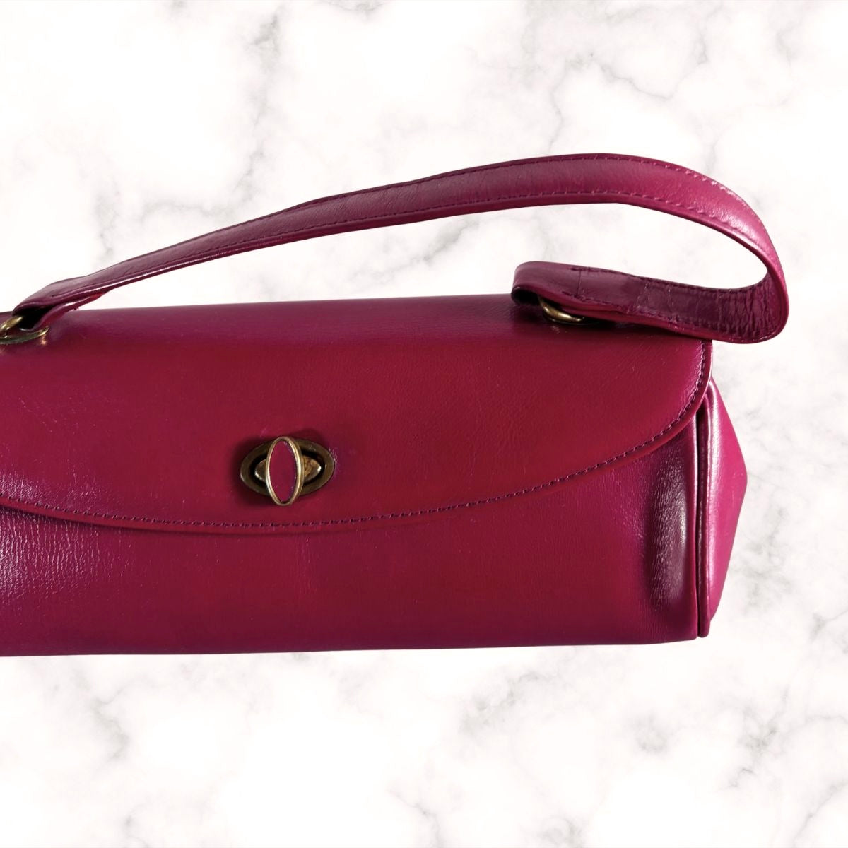 A dark red  leather handbag with a curved top handle and a gold-tone oval clasp, shown against a white marble background.