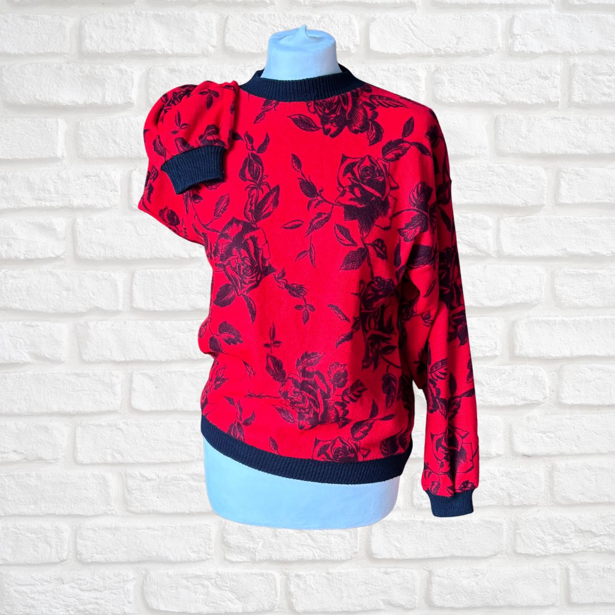 A red long-sleeve knit sweater with a black rose floral pattern, displayed on a light blue mannequin torso against a white brick wall background.