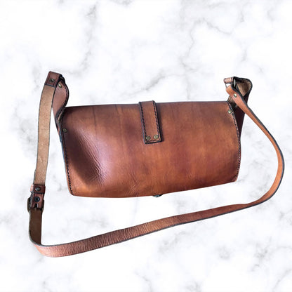 A brown leather shoulder bag with a long adjustable strap, laid flat on a white marble background.