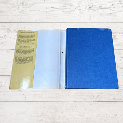 An open book  lying flat on a light whitewashed wood surface. The right page has a textured royal blue cover, and the left page is a tan/gold page with black text under a clear plastic sleeve.