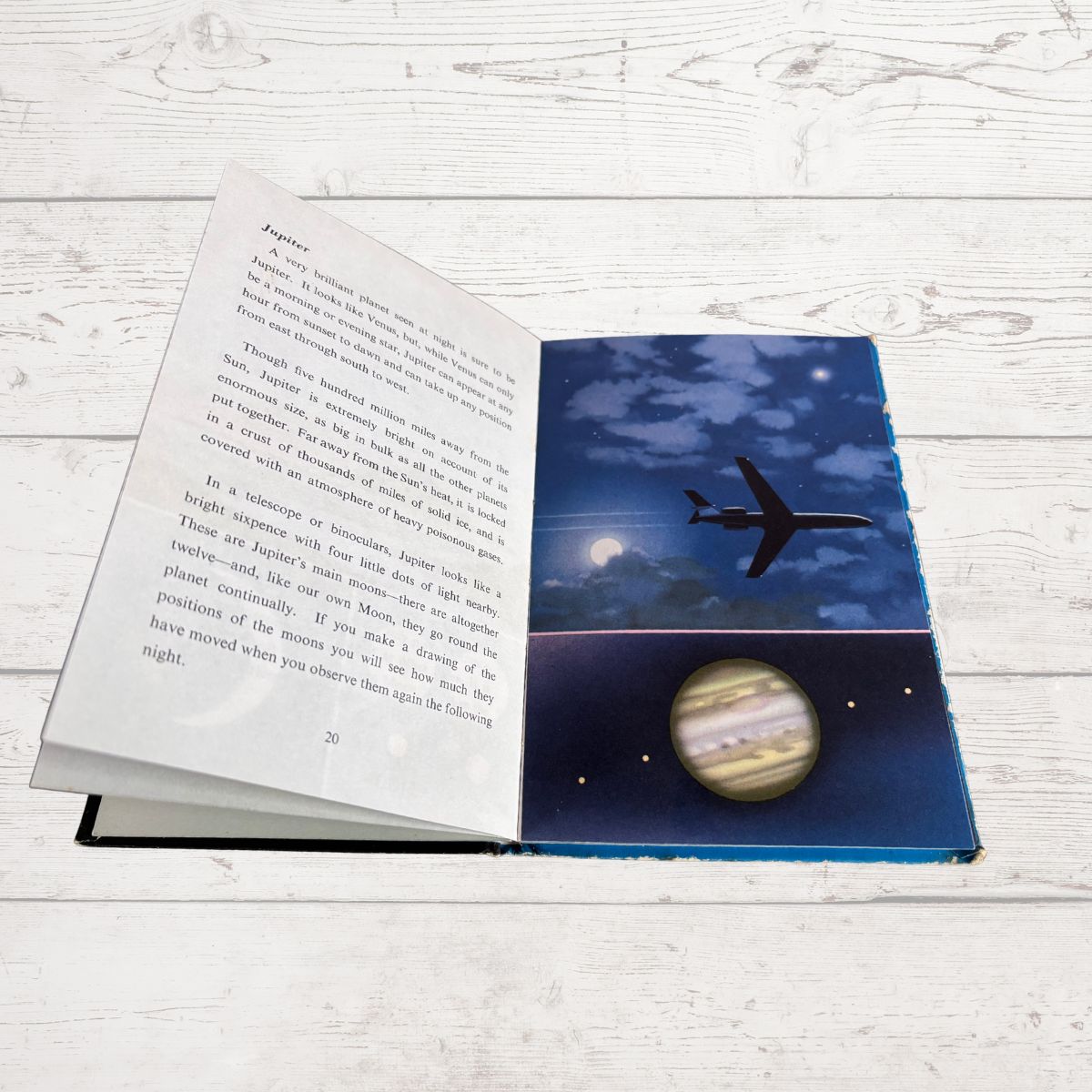 An open children's book lying on a white wood surface, showing printed text on the left page and a colorful space-themed illustration on the right page with a blue night sky, moon, stars, an airplane, and a planet.