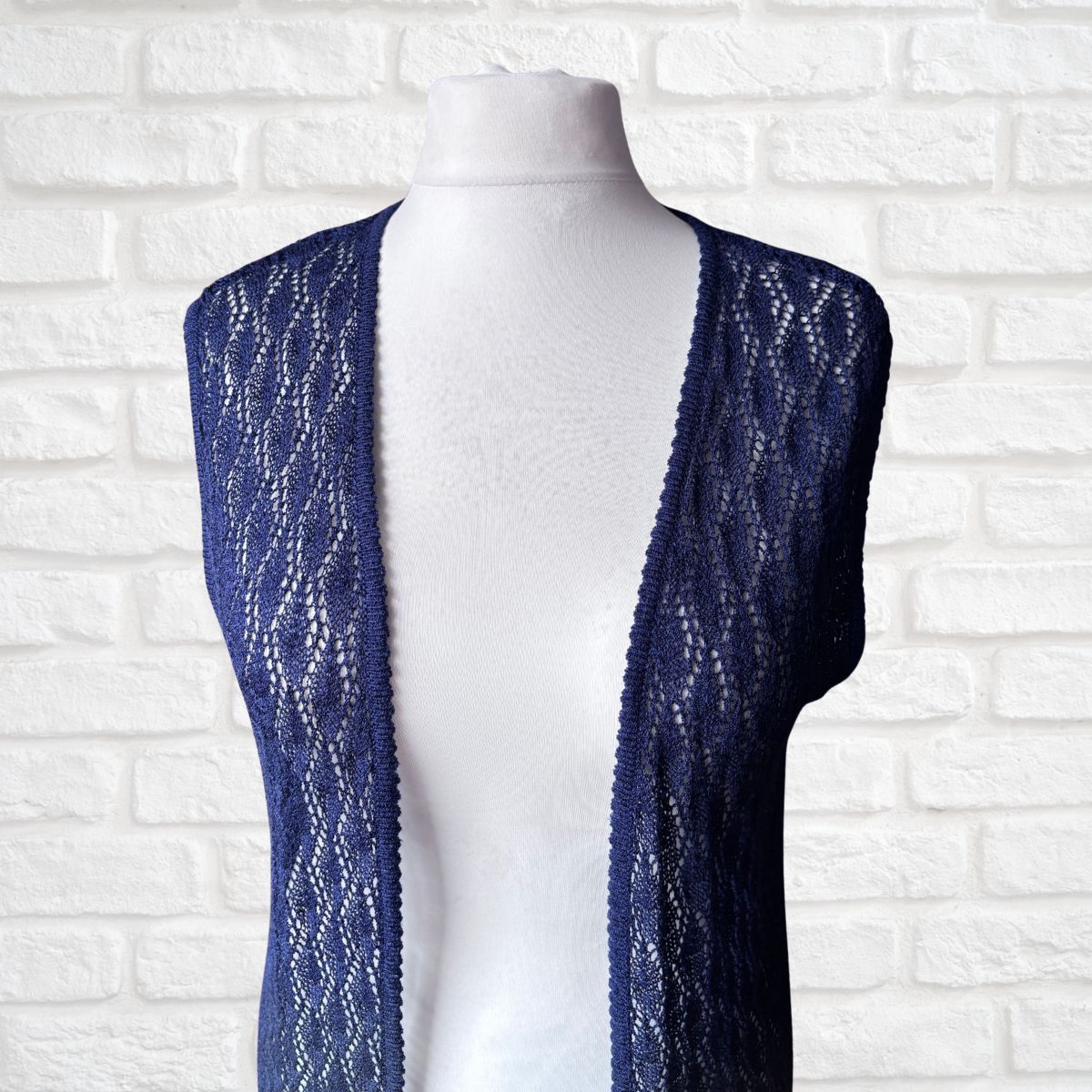 A navy blue open-front crochet knit vest displayed on a white mannequin torso against a white brick wall background.