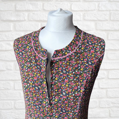 A sleeveless floral dress displayed on a white mannequin torso against a white brick wall background. The top features a dense multicolor floral pattern with pink, yellow, and white flowers on a dark base, with light pink piping trim around the collar and front opening.
