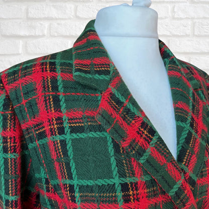 A close-up of a green plaid wrap jacket displayed on a white mannequin torso against a white brick wall background.