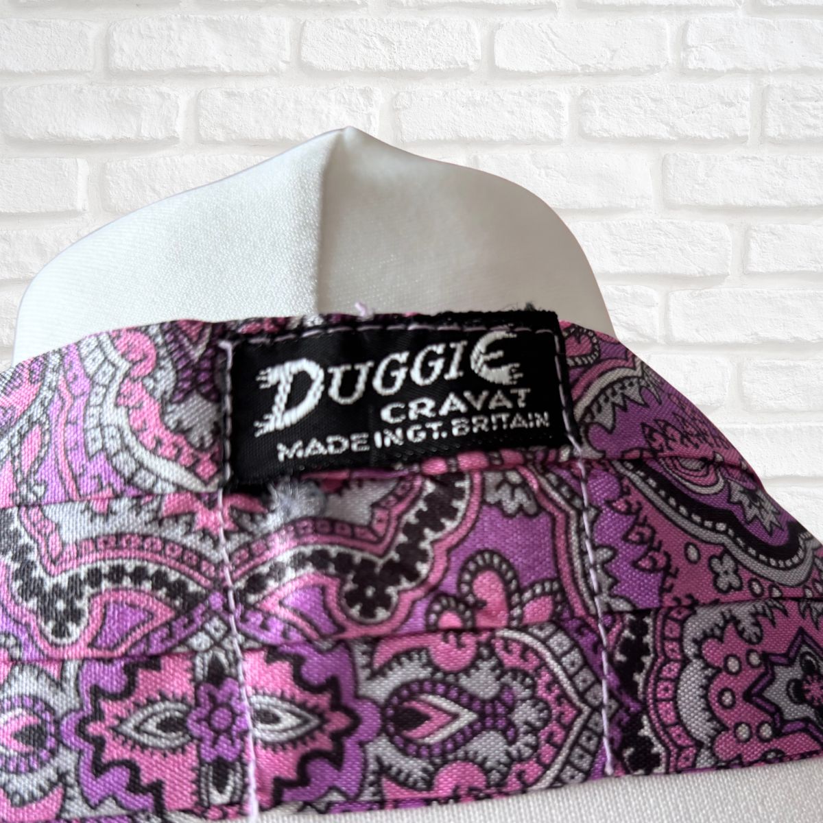 Purple paisley patterned cravat with 'Duggie Cravat' label on a white brick wall background