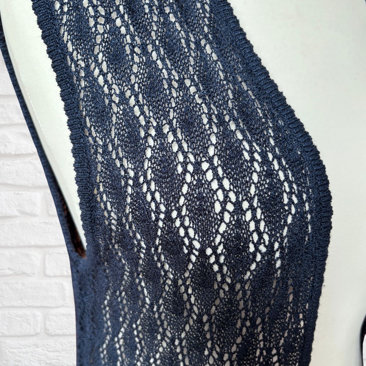 A close-up of a navy blue open-knit lace top or vest displayed on a white mannequin torso against a white brick wall background.