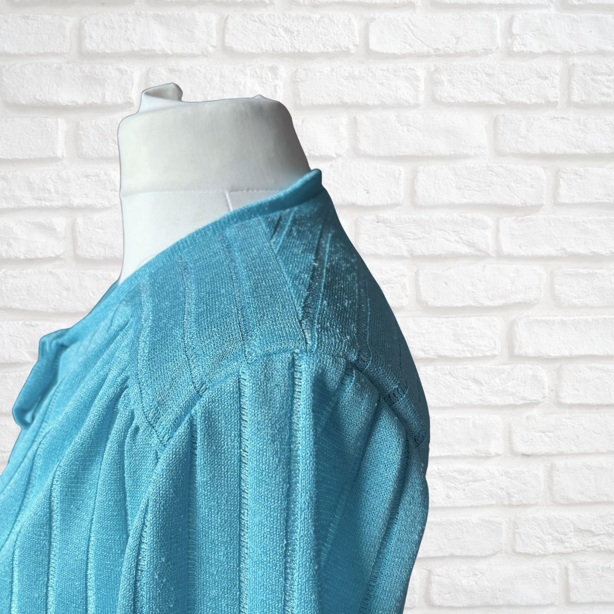 A close-up side view of a light blue knit jacket displayed on a white mannequin torso against a white brick wall background.