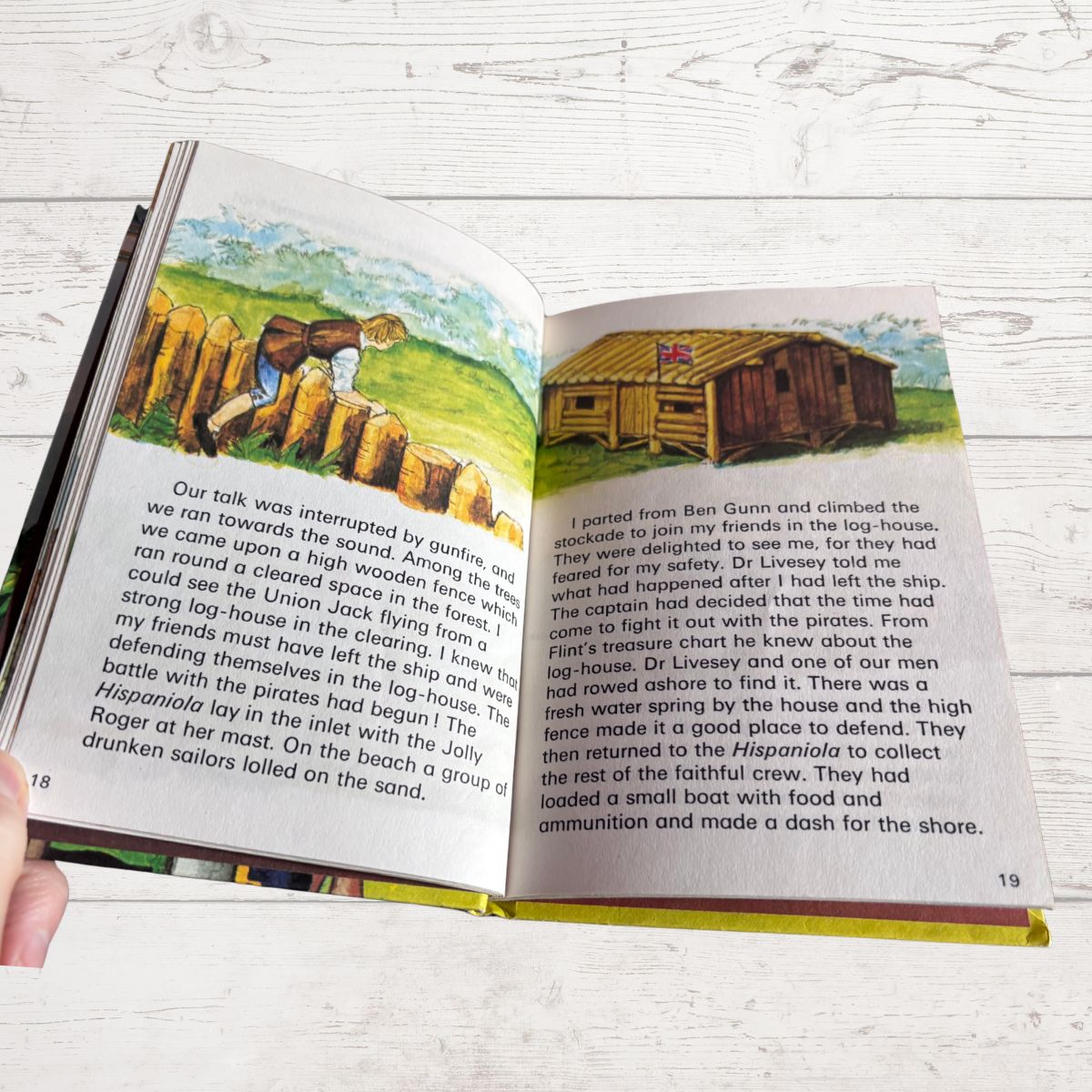 An open printed book spread showing two pages with text and a small inset photograph of a ship on the right page, held in a person's hand against a light wood background.