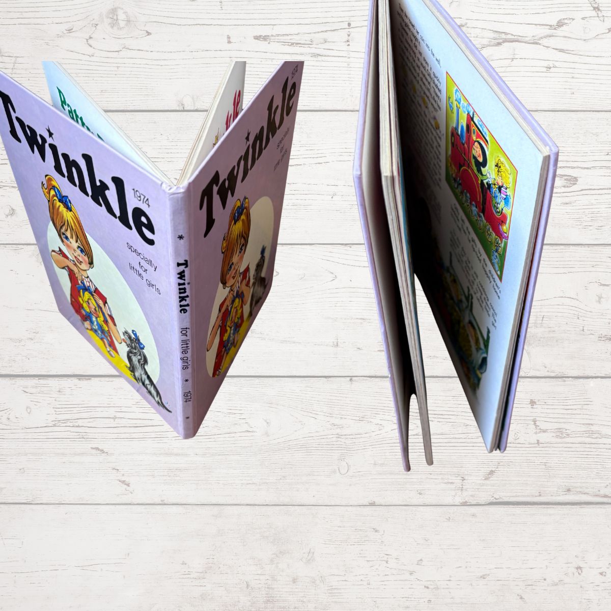 A children's book titled 'Twinkle' shown open in two views on a light wood plank surface. The cover is lavender with a cartoon illustration of a girl in a blue and yellow dress, and the interior pages contain colorful printed illustrations and text.
