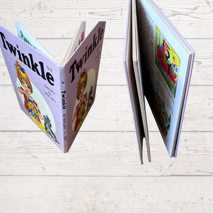 A children's book titled 'Twinkle' shown open in two views on a light wood plank surface. The cover is lavender with a cartoon illustration of a girl in a blue and yellow dress, and the interior pages contain colorful printed illustrations and text.