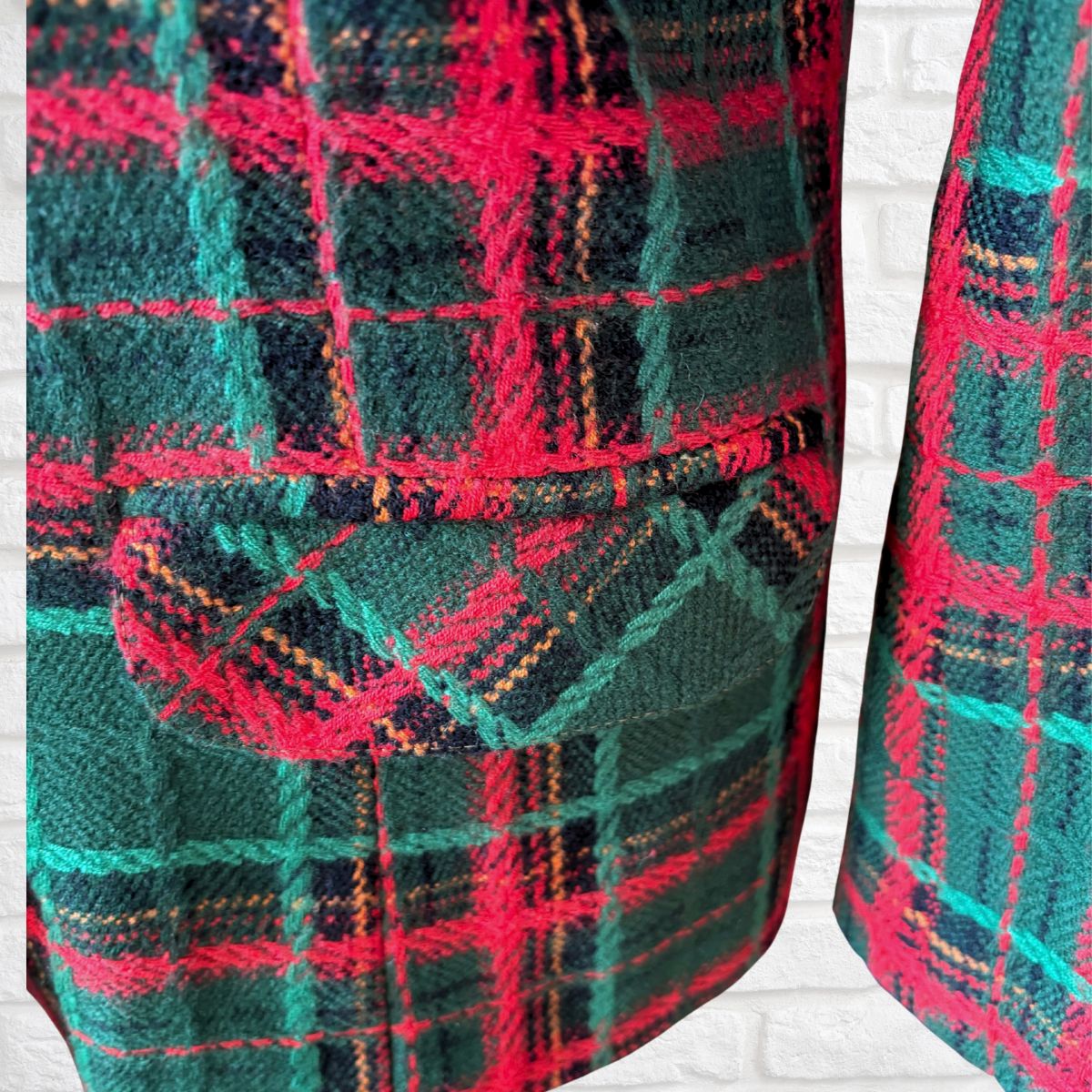 A close-up of a tartan plaid blazer featuring a pattern of dark green, black, and bright pink with thin orange and teal stripes, shown against a white brick wall background.