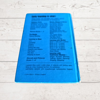 The back cover of a bright blue children's book titled 'Early Learning is vital ' lying flat on a white wood plank surface. The cover features black text detailing the publisher, Lambton Books, and learning topics covered.