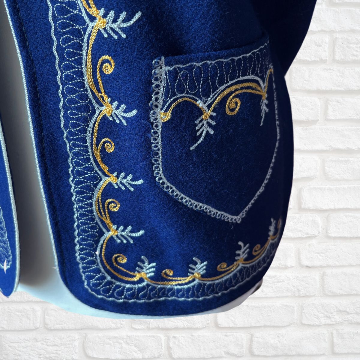 Blue waistcoat  with intricate white and gold embroidery on a white brick wall background