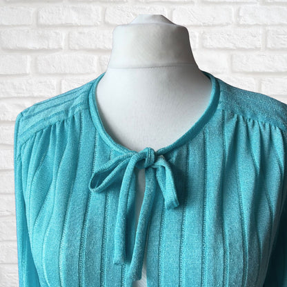 A turquoise blue ribbed knit cardigan with a tie-neck detail, displayed on a white mannequin torso against a white brick wall background.