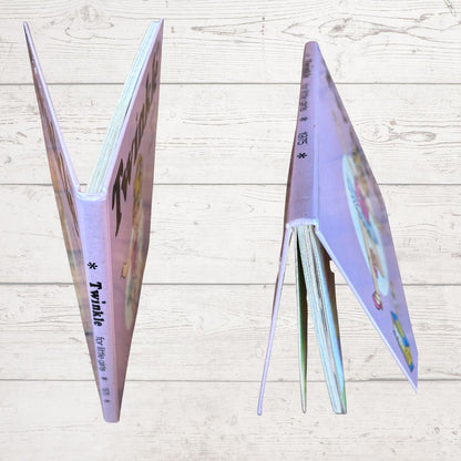 A children's picture book titled 'Twinkle' shown standing open in two views against a light wood plank background. The book has a lavender-purple cover with colorful illustrations and the title 'Twinkle' printed on the spine.