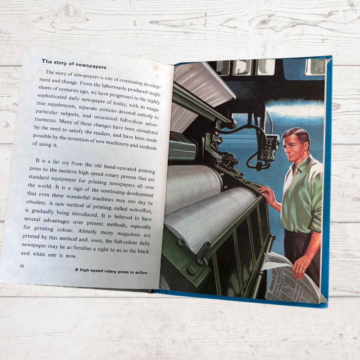 An open book lying on a light wood surface, showing printed text on one page and a colorful illustration on the other. The illustration depicts a man in a blue suit standing beside a large printing press and holding a newspaper.