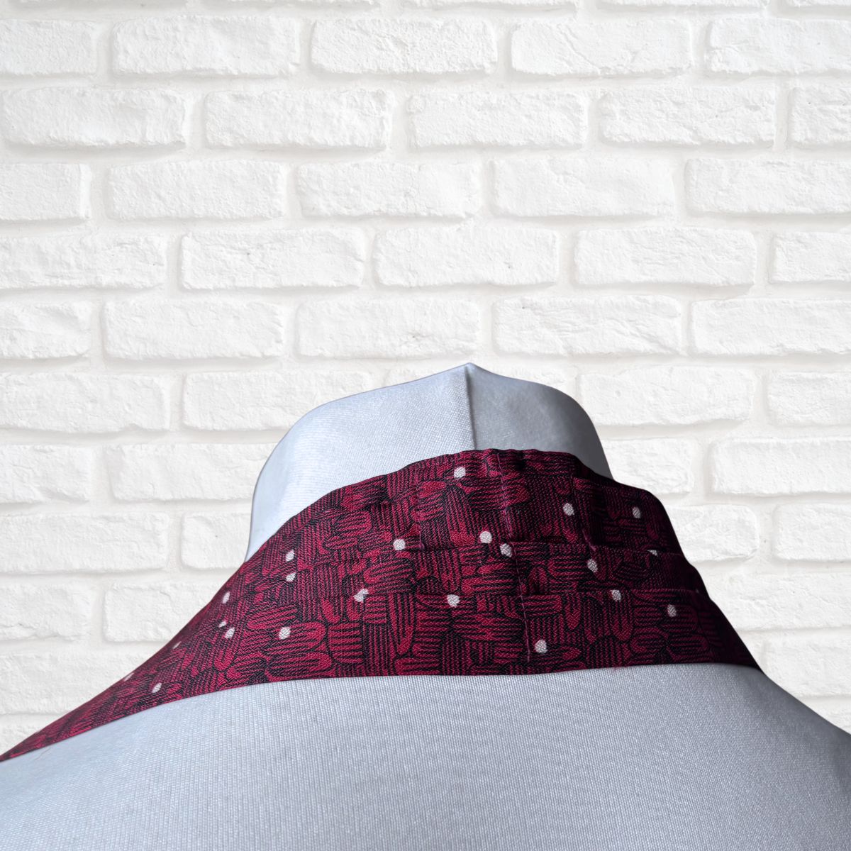 Mannequin with a burgundy patterned cravat against a white brick wall background