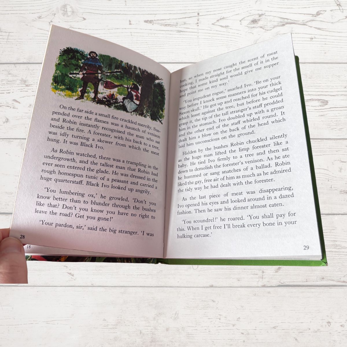 An open children's book being held by a hand, showing printed text and a black-and-white illustration on both pages against a light wood background.