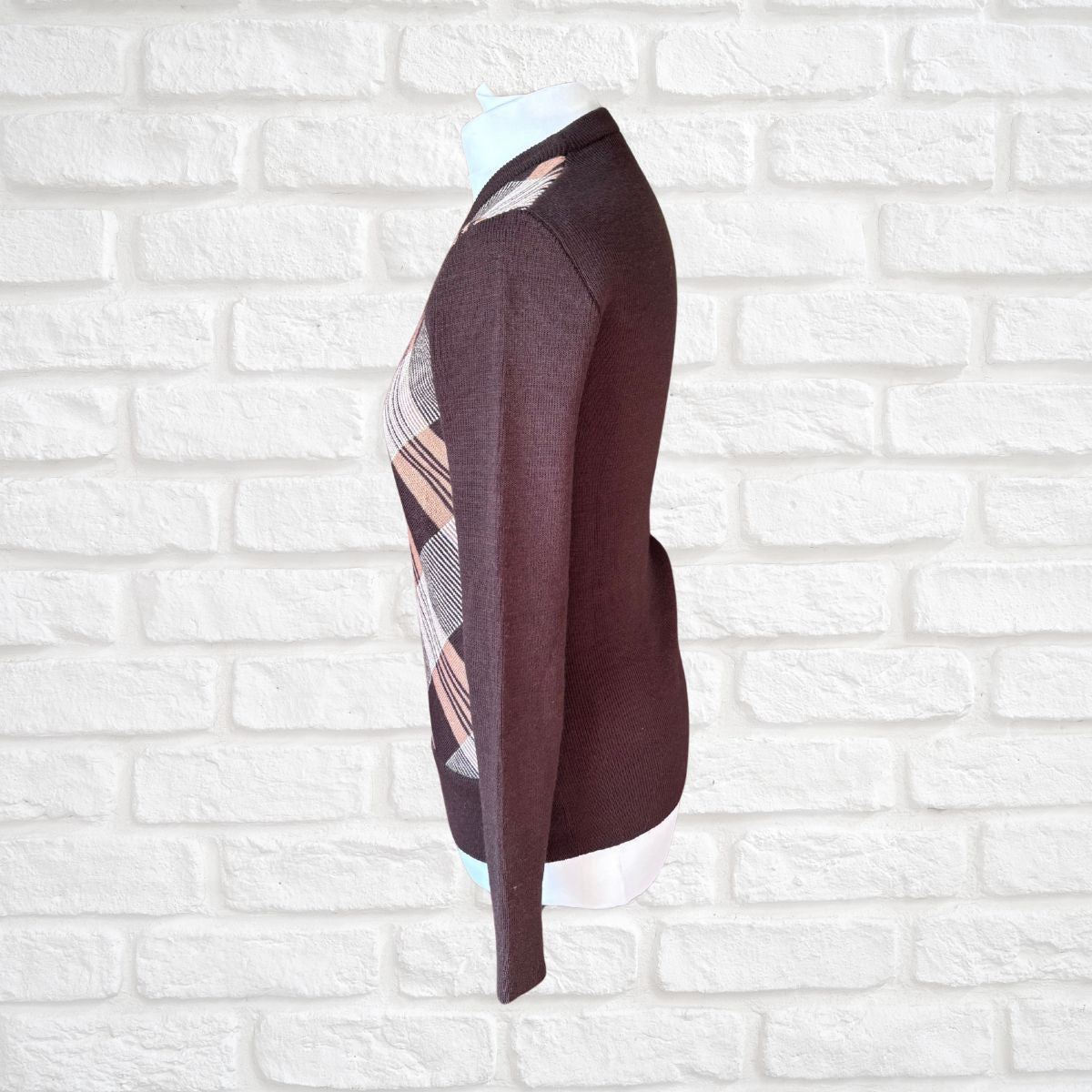 A dark brown knit  sweater with a beige and white  plaid pattern panel on the side, displayed on a white mannequin torso against a white brick wall background.