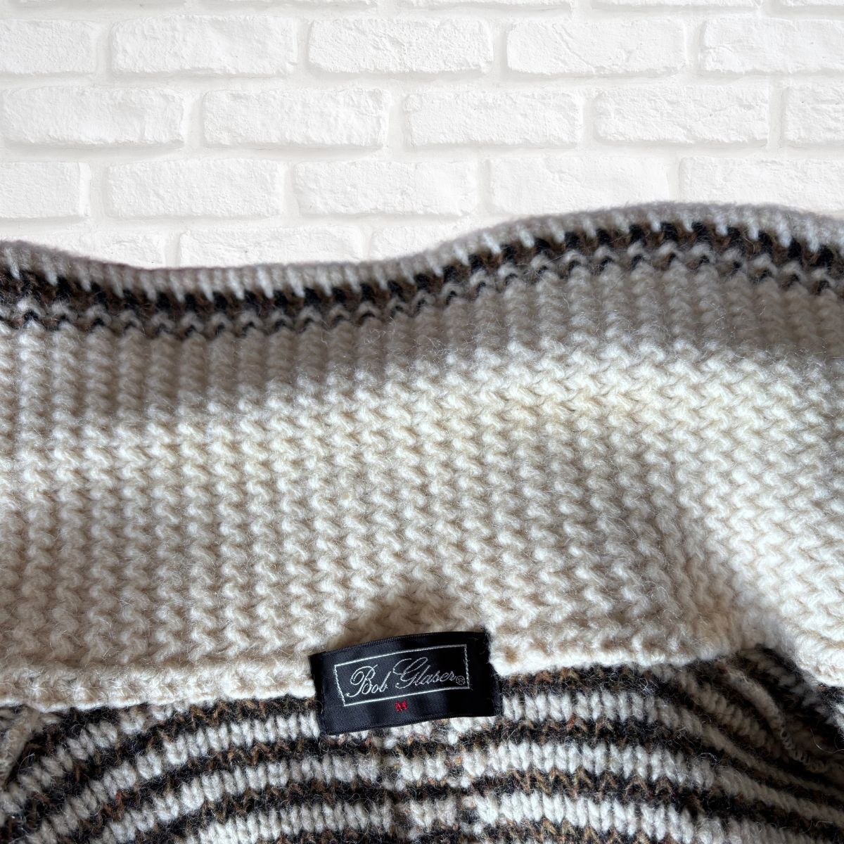 A close-up of a knitted cardigan with a cream and dark brown striped pattern, featuring a black fabric label that reads 'Bed Collection'. The blanket is draped against a white painted brick wall background.