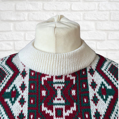 A close-up of a knit sweater on a cream-colored dress form, featuring a red, green, and white geometric pattern with a cream ribbed turtleneck, set against a white brick wall background.