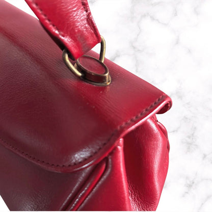 A close-up of a red leather handbag showing the flap closure, stitching, and a brass-tone metal ring attachment, set against a white marble background.