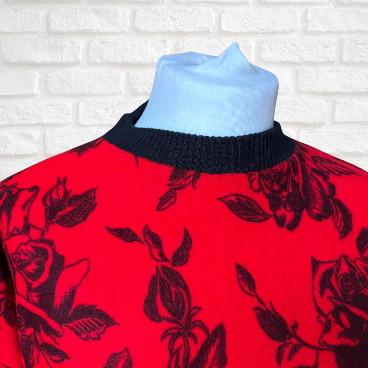 A red knit sweater with a black rose and leaf pattern, displayed on a light blue mannequin torso against a white brick wall background.