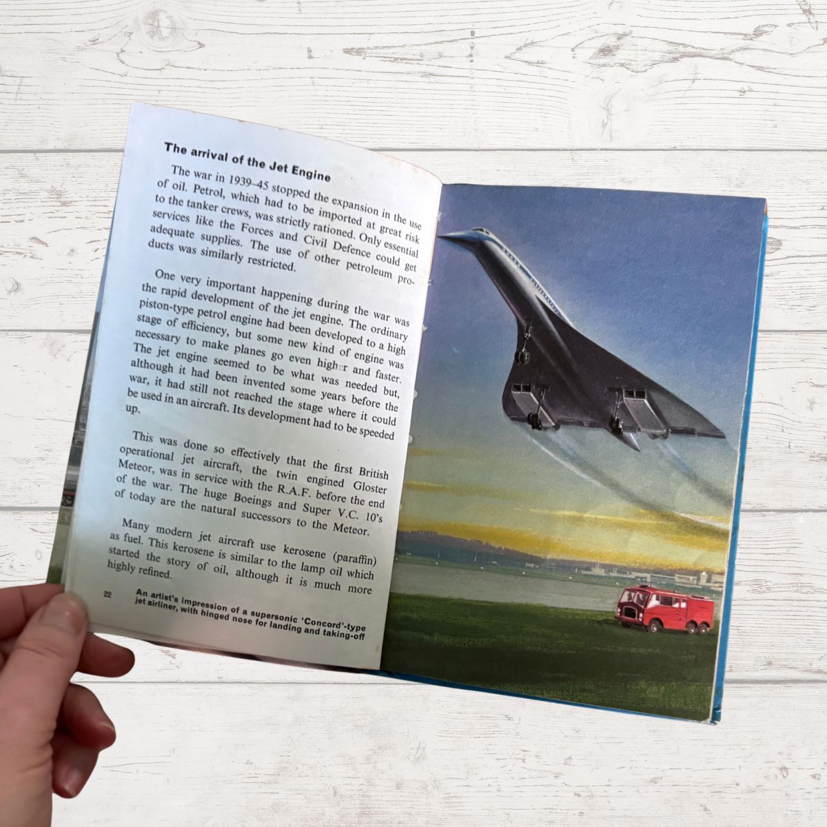 An open children's book lying on a white wood surface, showing a printed page about jet engines and a large color illustration of a jet aircraft over a green field with a red fire truck. A human hand is visible holding the left edge of the book.