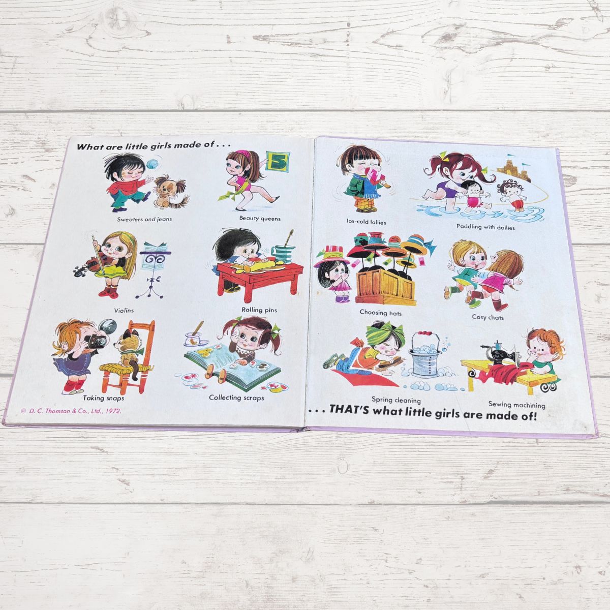An open children's book laid flat on a light wood surface. The pages contain cartoon illustrations of children performing various activities, with text such as 'What are little girls made of.