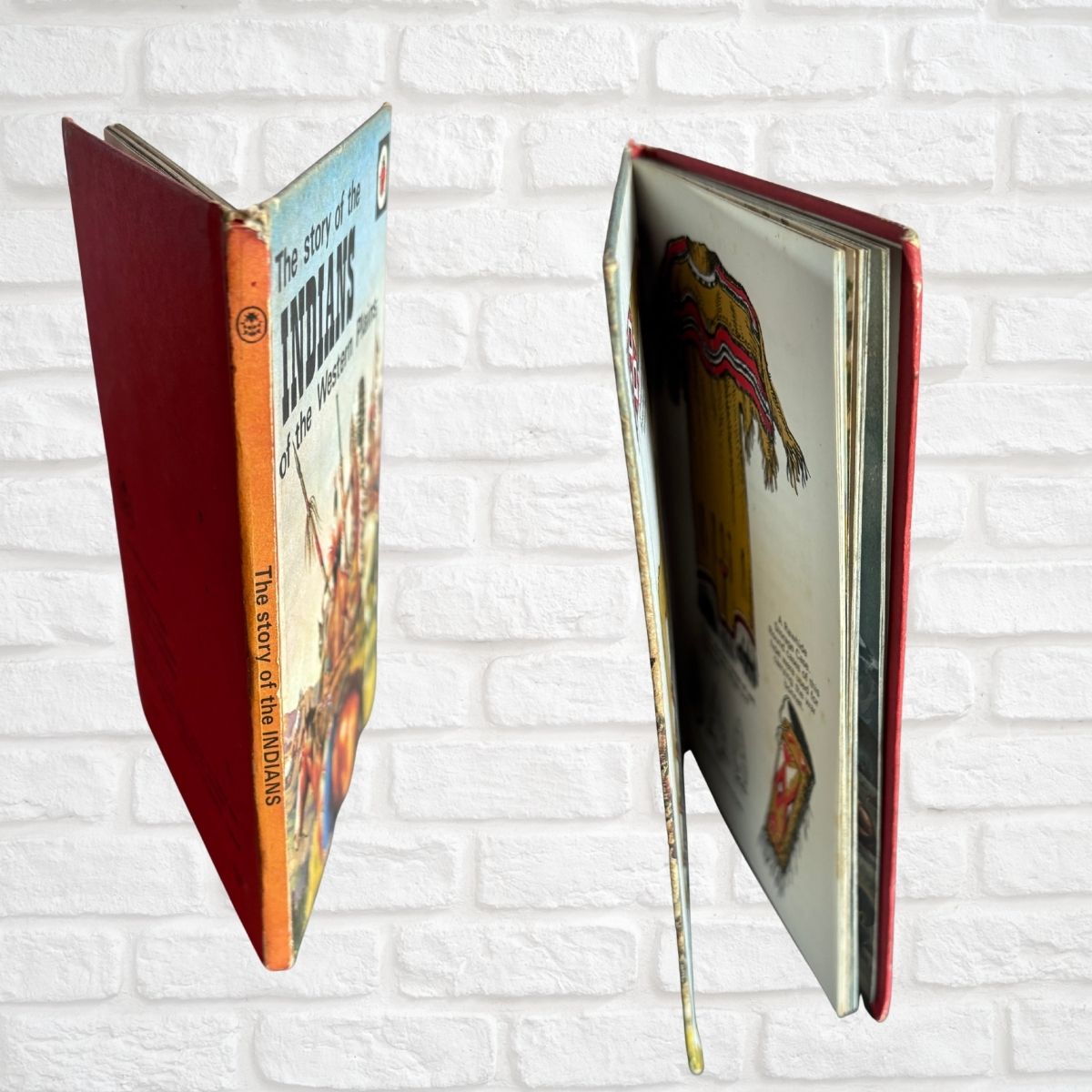 A vintage hardcover book shown from two angles against a white brick wall. One view shows the red spine with the title 'The story of the Koolaus' and an orange band, while the other shows the interior pages with illustrations.