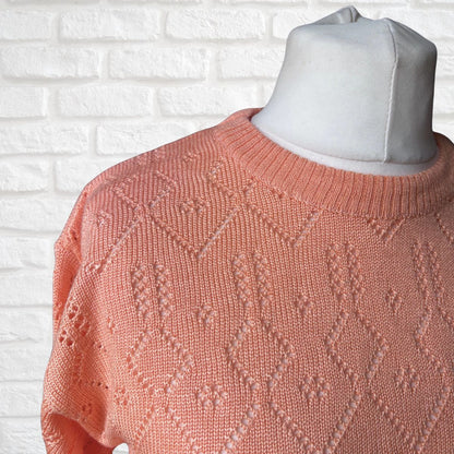 A close-up of a peach-colored knit sweater with a textured geometric pattern, displayed on a light gray mannequin torso against a white brick wall background.