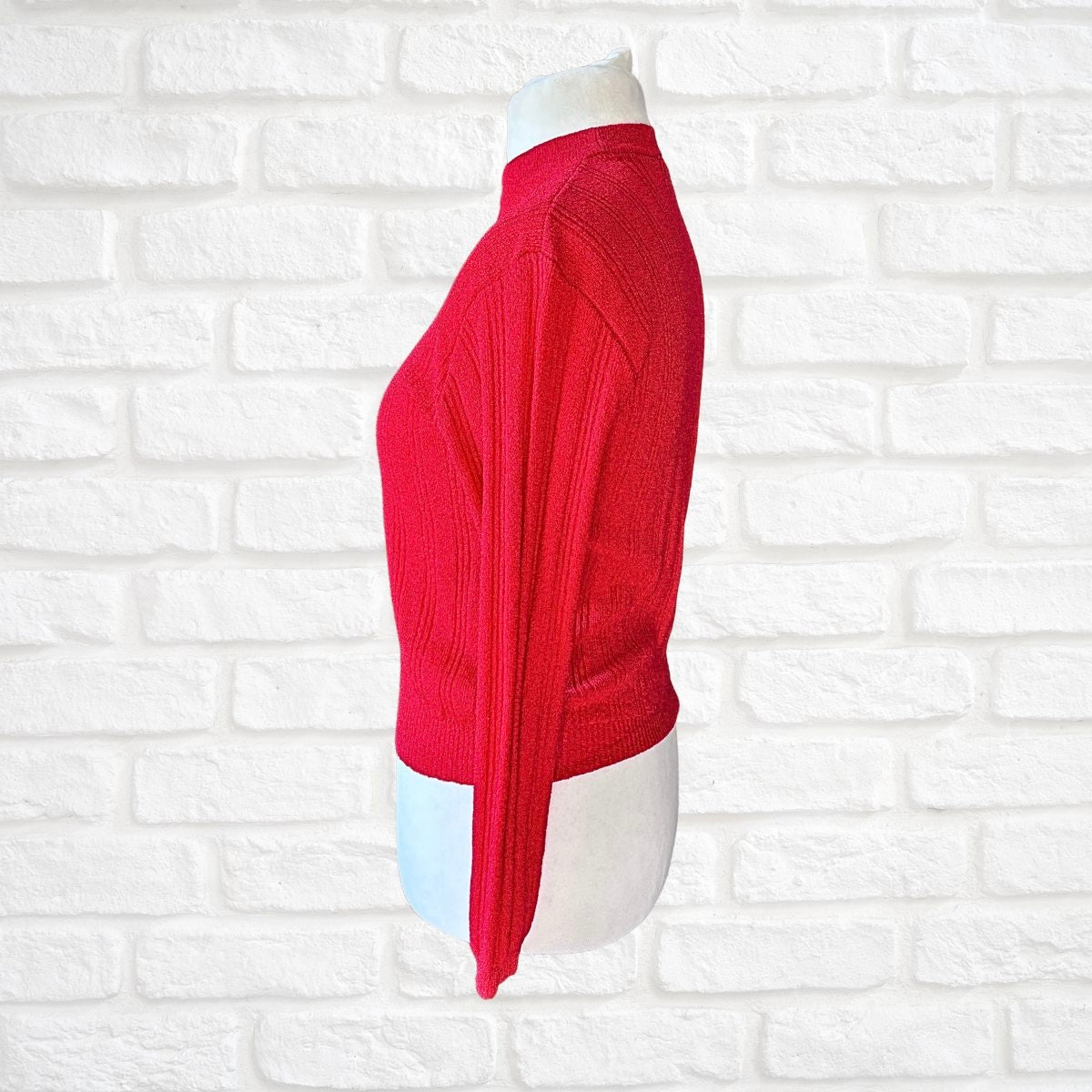 A bright red ribbed knit mock-neck sweater displayed on a white mannequin torso, set against a white painted brick wall background.