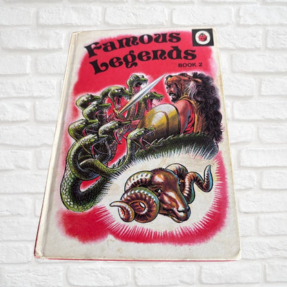 Vintage book titled 'Famous Legends Book 2' with a red cover illustration featuring a lion, snakes, and a ram, photographed against a white brick wall.