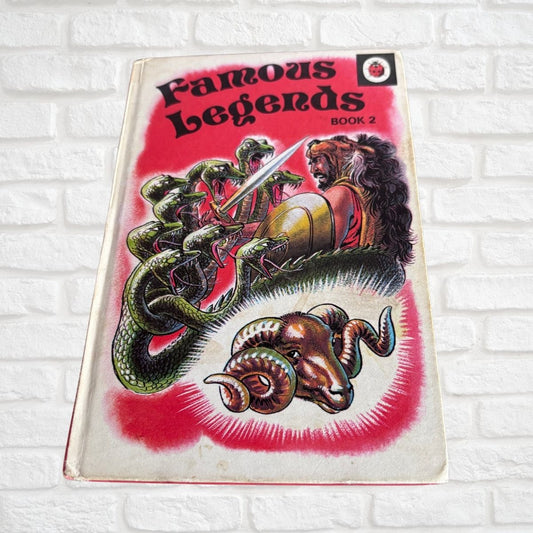 Vintage book titled 'Famous Legends Book 2' with a red cover illustration featuring a lion, snakes, and a ram, photographed against a white brick wall.