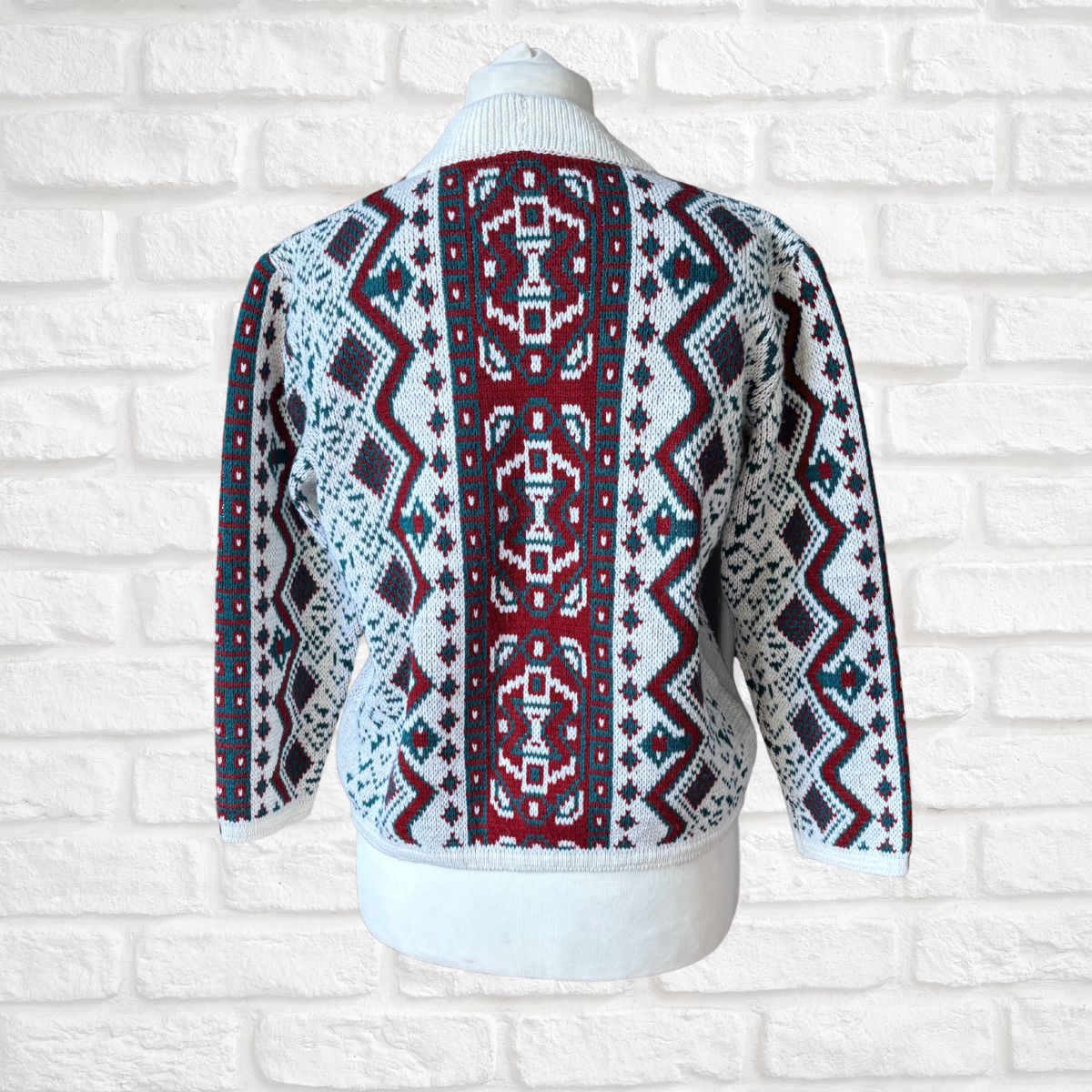 A knit sweater with a complex geometric and tribal-inspired pattern, displayed on a white mannequin torso against a white brick wall background.