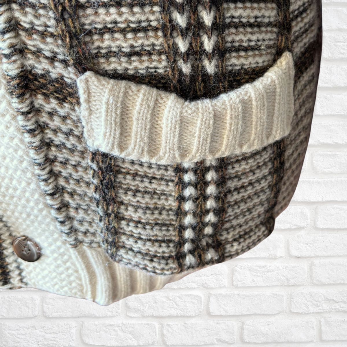 Close-up of a chunky knit cardigan featuring a textured plaid pattern in cream, brown, and black, with a ribbed cream pocket and a silver button, set against a white brick wall background.