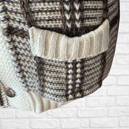 Close-up of a chunky knit cardigan featuring a textured plaid pattern in cream, brown, and black, with a ribbed cream pocket and a silver button, set against a white brick wall background.