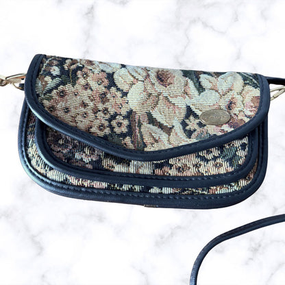 A vintage-style floral tapestry crossbody bag with navy blue trim, resting on a white marble surface. The bag features a detailed woven floral pattern in beige, brown, and muted pink tones, with a small oval metal logo plate on the front flap.