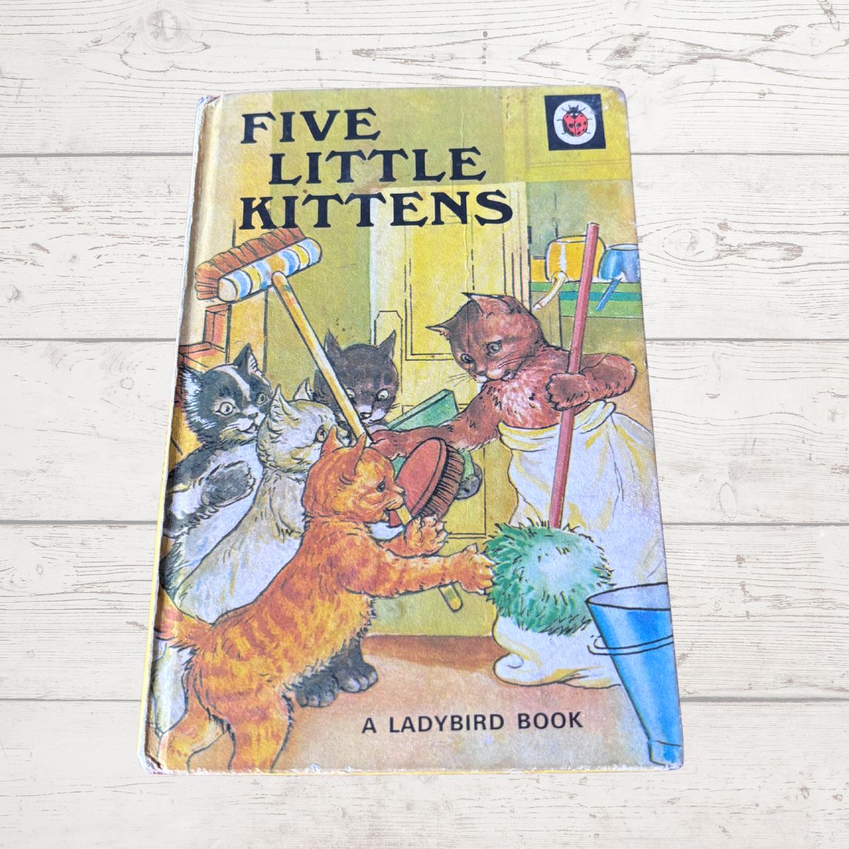 Vintage children's book titled 'Five Little Kittens' by Ladybird, photographed on a light wood surface. The cover shows five illustrated kittens cleaning up with toys and a dustpan.