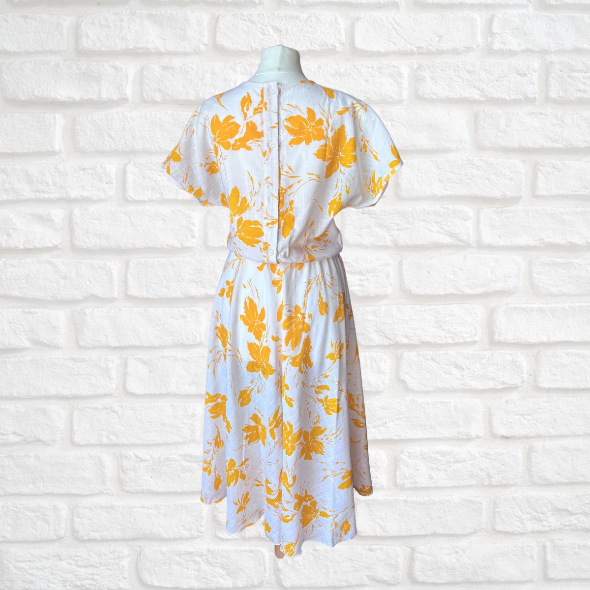 White dress with yellow floral pattern on a mannequin against a white brick wall.