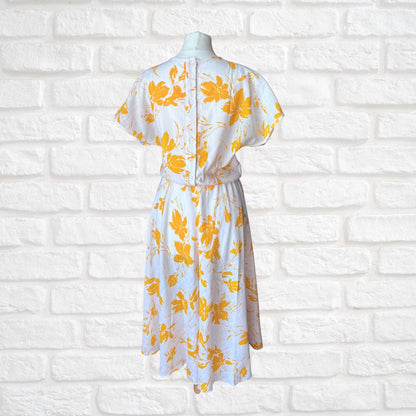 White dress with yellow floral pattern on a mannequin against a white brick wall.