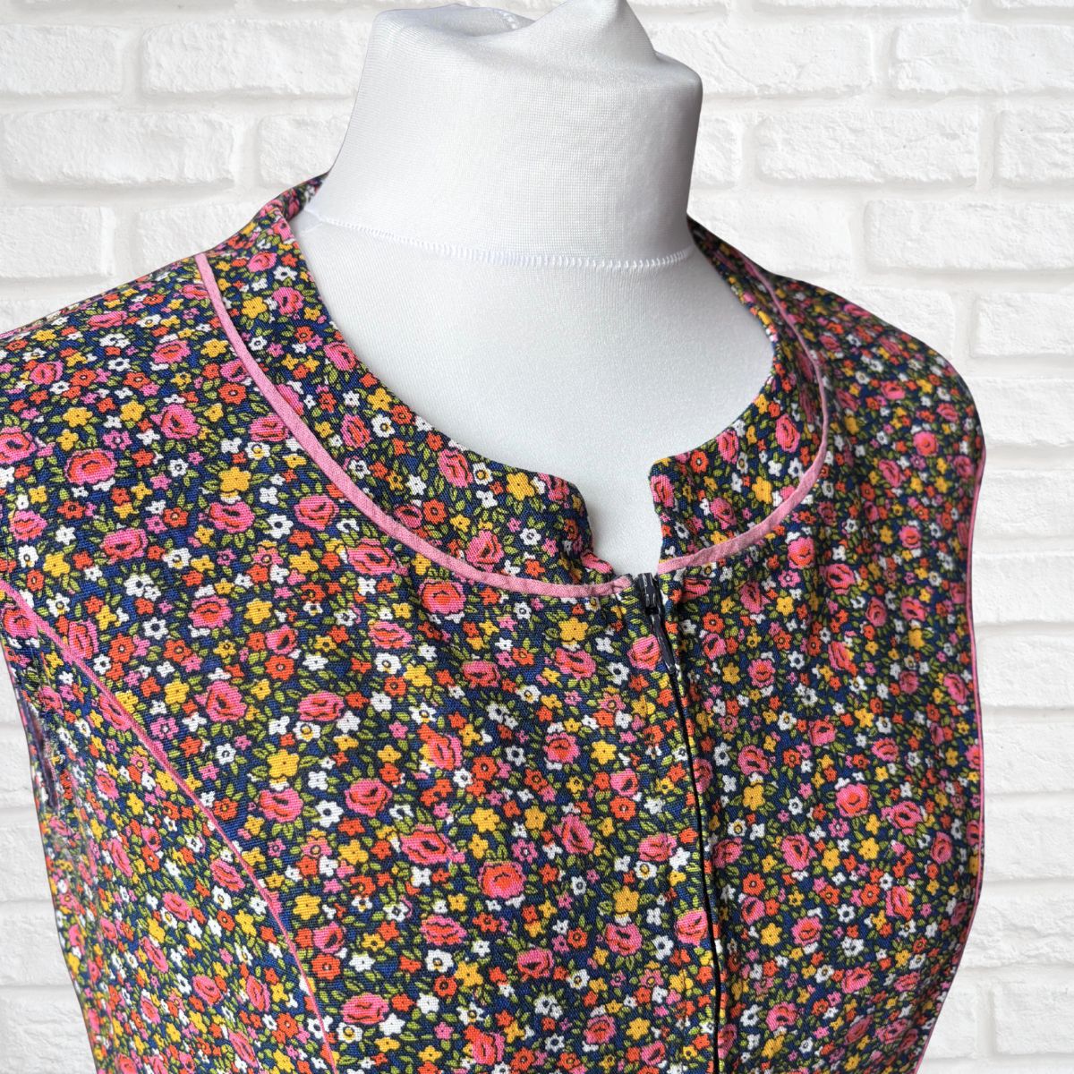 A sleeveless dress with a dense multicolour floral print on a dark background, featuring a pink-trimmed split neckline and a front zipper, displayed on a white mannequin torso against a white brick wall.
